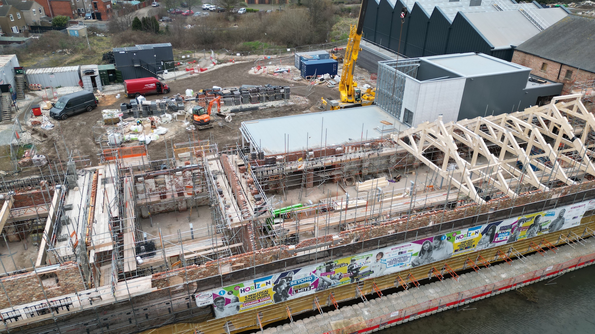 At the construction site, partially built structures rise alongside a large yellow crane, signaling contributions to Grimsby's Future. Nearby, assorted building materials and machinery surround a red vehicle and orange equipment, setting the foundation for the Horizon Youth Zone.